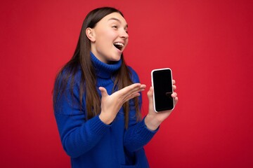 Photo of pretty happy young brunette woman wearing casual blue sweater isolated over red background with empty space holding in hand mobile phone and showing smartphone with empty screen for mockup