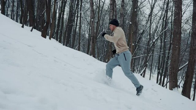 Slow motion man in sportswear, hat and gloves running up the hill covered with snow, trees on background. Tracking shot of athlete training in forest in winter. Concept of sport