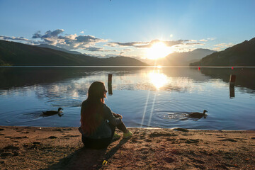 A woman sitting at the shore of Millstaetter lake during the sunset. High Alps around. The sun sets behind thick clouds. Calm surface of the lake reflects the sky. A swan and a duck swinging around. - Powered by Adobe