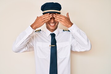 Young hispanic man wearing airplane pilot uniform covering eyes with hands smiling cheerful and funny. blind concept.
