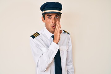 Young hispanic man wearing airplane pilot uniform hand on mouth telling secret rumor, whispering malicious talk conversation