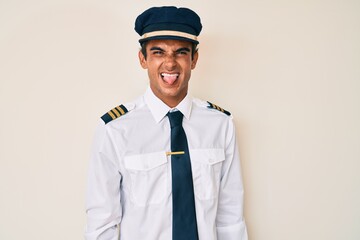 Young hispanic man wearing airplane pilot uniform sticking tongue out happy with funny expression. emotion concept.