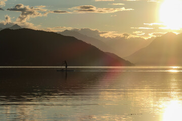 A man stand up paddling during the sunset on Millstaetter lake in Austria. The lake is surrounded by high Alps. Calm surface of the lake reflecting the sunbeams. The sun sets behind the mountains.