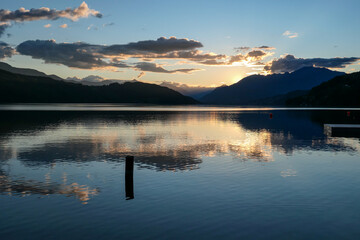 A sunset by Millstaetter lake in Austria. The lake is surrounded by high Alps. Calm surface of the lake reflecting the sunbeams. The sun sets behind the mountains. A bit of overcast. Natural beauty