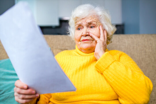 silver-haired mature russian unhappy woman holding at banking paper bill in living room at modern house
