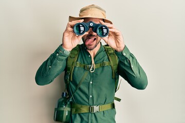 Handsome man with beard wearing explorer hat looking through binoculars sticking tongue out happy with funny expression.