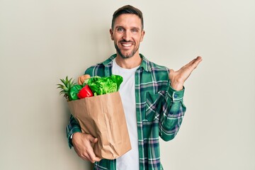 Handsome man with beard holding paper bag with groceries smiling cheerful presenting and pointing with palm of hand looking at the camera.