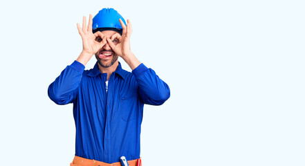 Young handsome man wearing worker uniform and hardhat doing ok gesture like binoculars sticking tongue out, eyes looking through fingers. crazy expression.