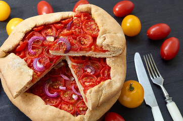 Sliced tomato galette or tart with onion rings on the dark wooden board decorated with various cherry tomatoes. Traditional French vegetarian food on the black background