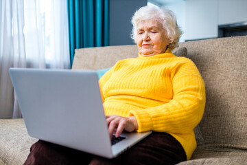 aged mature grey haired woman in yellow sweater sitting on sofa with laptop in living room