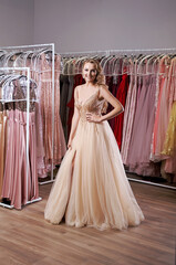 Young beautiful blonde girl wearing a full-length beige yellow champaign chiffon slit prom ball gown decorated with golden sparkles and sequins. Fashion model in dress hire service with many dresses.