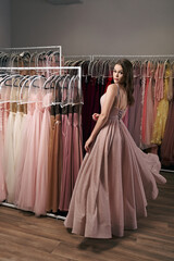 Portrait of young beautiful girl wearing a full-length pale pink glitter chiffon draped prom ball gown. Model selecting an outfit for occasion in dress hire service with many options on background.