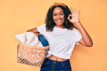 Young african american woman holding laundry basket doing ok sign with fingers, smiling friendly...