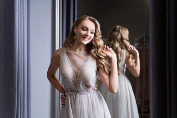 Young beautiful blonde girl wearing a full-length silver white chiffon prom ball gown decorated with sparkles and sequins. Model in front of mirror in a fitting room at dress hire service. © berezko