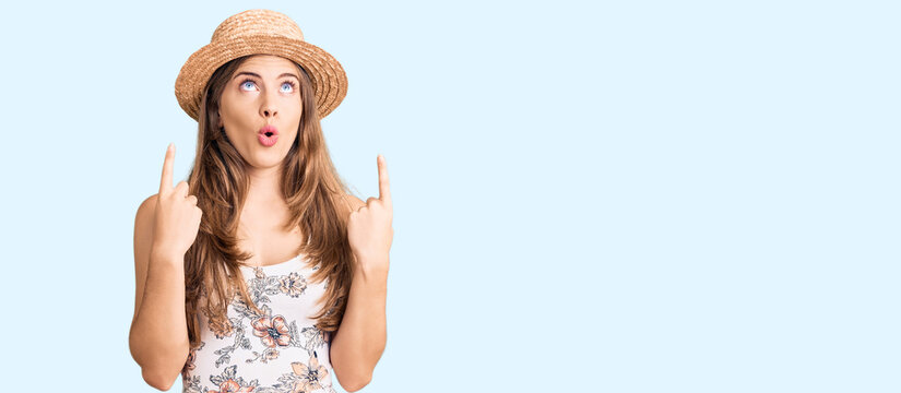 Beautiful caucasian young woman wearing summer hat amazed and surprised looking up and pointing with fingers and raised arms.