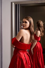 Young beautiful girl wearing an off-the-shoulder full-length crimson red satin slit prom ball gown. Model looking in mirror. Fitting room in dress hire service.