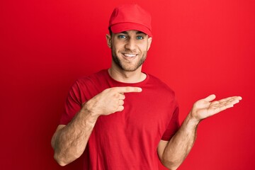 Young caucasian man wearing delivery uniform and cap amazed and smiling to the camera while presenting with hand and pointing with finger.