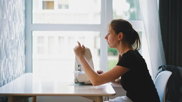side shot: young woman with long dark hair sits at white table in front of window during remote work, adjusting fabric and sewing machine
