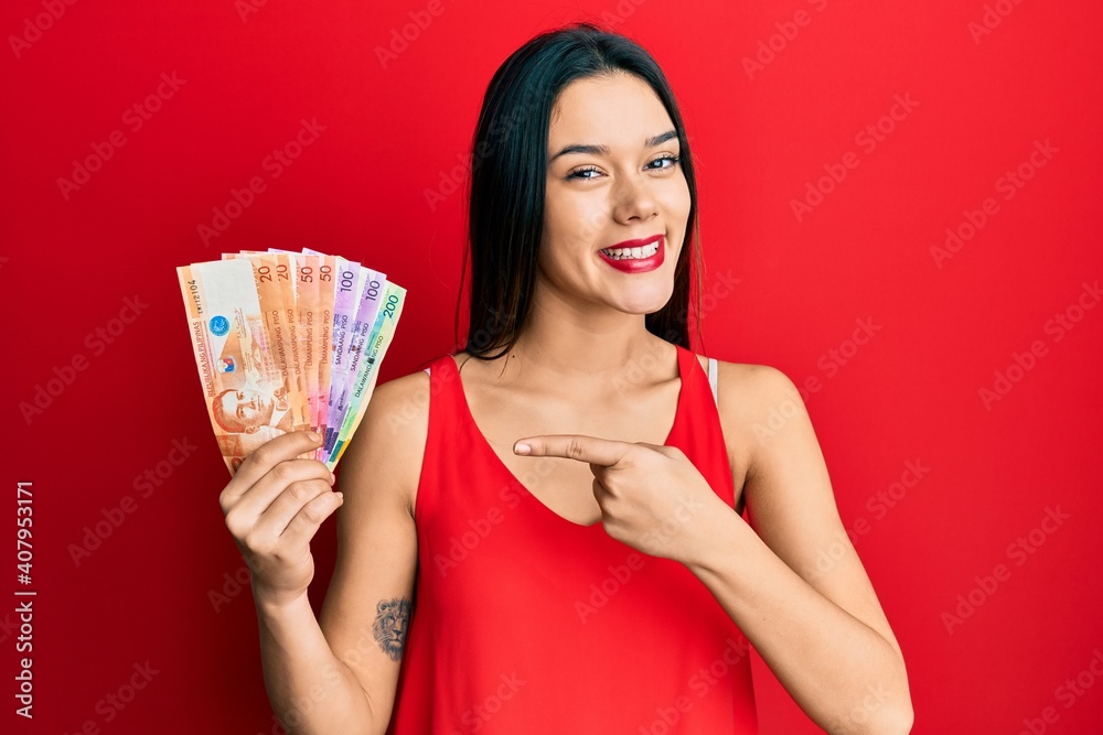 Wall mural Young hispanic girl holding philippine peso banknotes smiling happy pointing with hand and finger