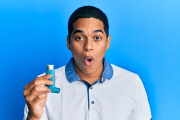 Young handsome hispanic man holding medical asthma inhaler scared and amazed with open mouth for...