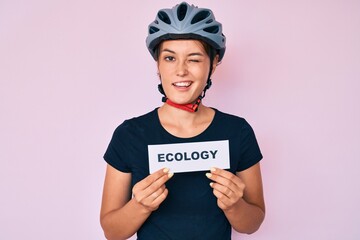 Beautiful caucasian woman wearing bike helmet holding ecology word winking looking at the camera...