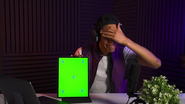 Multi-ethnic man watches in tablet and reacts emotionally. Chromakey green screen on the tab. Professional vlogger make content in his studio. Colored light on background