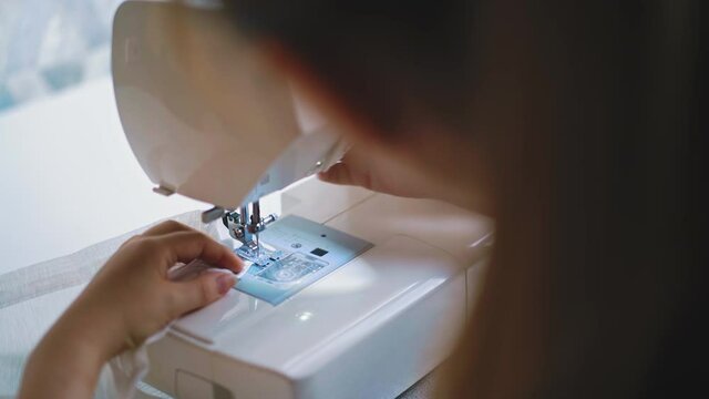 close up, back side view: young woman performs an order to sew curtains, at home, remote work, adjusts correct course of needle at white sewing machine sitting at table