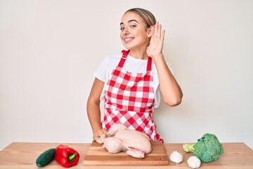 Young beautiful blonde woman wearing apron cooking chicken smiling with hand over ear listening and hearing to rumor or gossip. deafness concept.