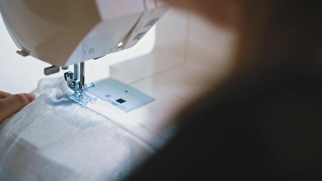 close view, back side shot: cute brunette woman sews edge of white transparent cloth, with help of electric sewing machine, holds her palms, take corner by her hand, then check