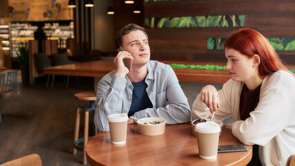 Two teenagers spending time together, sitting in a cafe on a daytime. Girl looking bored, eating her meal while her boyfriend is ignoring her, talking on the phone