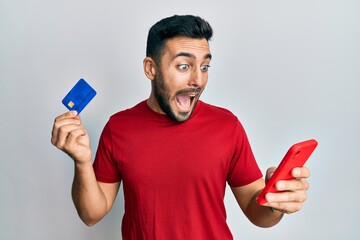Young hispanic man holding smartphone and credit card celebrating crazy and amazed for success with...