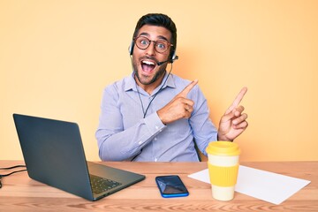 Young hispanic man sitting at the desk wearing operator headset at the call center office smiling and looking at the camera pointing with two hands and fingers to the side.