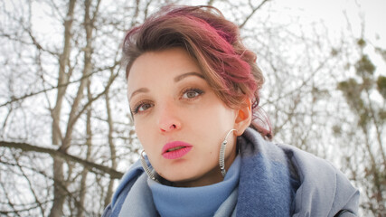 Attractive young woman with perfect skin and makeup with dark pink hair is possing on winter park background wearing blue scarf and silver earrings