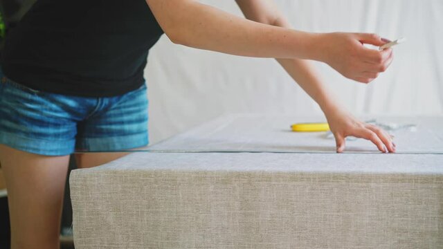 young slender woman in short shorts, at home during quarantine, engaged in cutting dense gray fabric, applies metal ruler, draws chalk, sunlight