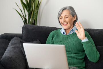 A smiling grandmother wearing a green jumper is sitting in the living room, relaxing and talking to her friend or family, waving at the laptop screen, staying connected during pandemic