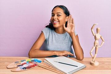 Young african american girl artist sitting at studio table smiling with hand over ear listening and hearing to rumor or gossip. deafness concept.