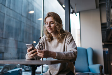 Focused woman using smartphone and holding glasses at cafe table
