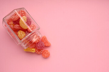 Sweet fruit hard candies pouring out of the glass mason jar on colored pink background. Overhead view of colorful caramel assortment.