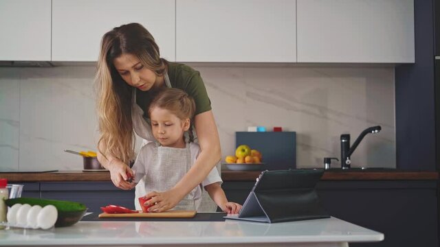 young sweet mother in gray apron stands in kitchen, at table with tablet computer and ingredients in plates, trains little daughter to properly hold knife in her hand and cut vegetables