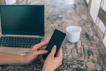 Freelancer using smartphone while sitting at wooden table with laptop