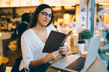 Positive caucasian female in eyewear for vision correction enjoying reaming to do list and plannings working remote in cafeteria, smiling woman clogger checking publications and articles on work