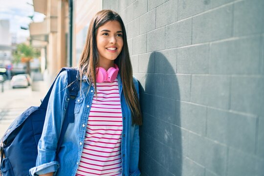 Young hispanic student girl smiling happy using headphones leaning on the wall at the city.
