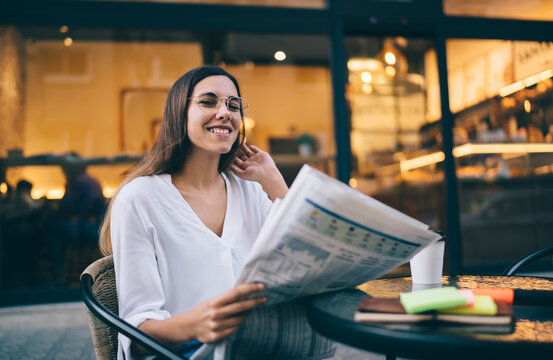 Portrait Of Cheerful Caucasian Female In Eyewear For Vision Correction Holding Newspaper Spending Coffee Break On Cafe Terrace, Smiling Woman 20s Searching Information And Vacancy From Printed Paper