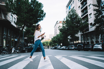 Smiling caucasian female in casual wear walking on zebra in city holding mobile phone with navigation application, positive woman 20s millennial crossing road using smartphone unattentive in downtown