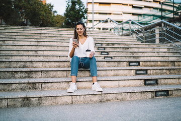 Young lady sitting on street staircase with smartphone and cup of coffee