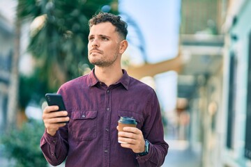 Young hispanic man with serious expression using smartphone and drinking coffee at the city.