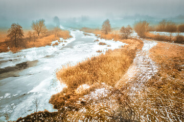 Thawed patches on the ice of a frozen river - landscape during the off-season in foggy weather. Melting river among yellowed grass and bare trees.