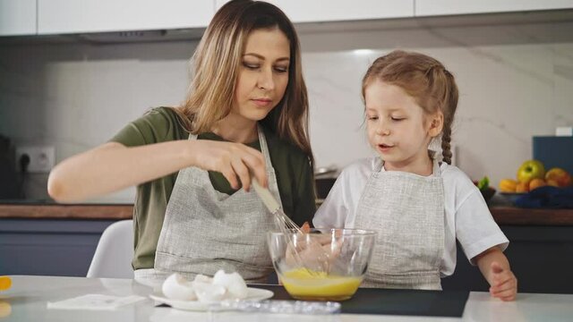 moving background: young mother shows her little daughter in kitchen how to beat eggs in deep bowl with whisk, girl tries to beat eggs, winces from tension