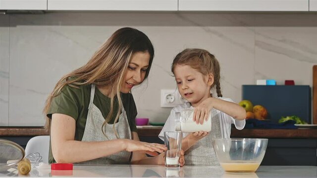 slow motion: young mother and daughter cook together food, girl pours flour into measuring transparent glass, accidentally hits mother on wrist, smiles on moving background