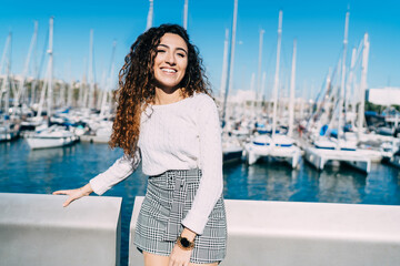 Half length portrait of cheerful hipster girl in trendy clothing satisfied with recreation time in quayside, happy Turkish woman smiling at camera while posing at pier with wharf berth on background